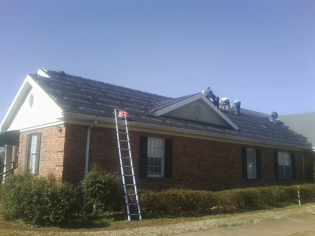 Crew tearing shingles off a brick ranch roof with a ladder set — Emergency Roof Repair work by our crew in Hallstead