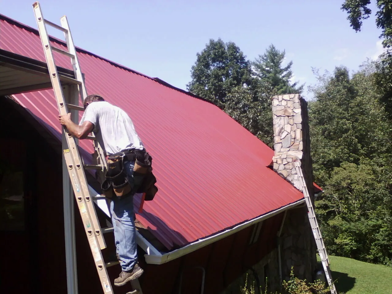 Roofer on a ladder beside a red metal roof and stone chimney — Soffit & Fascia Repair by Roofing Brothers in Hallstead